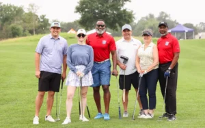 Six golfers standing together on the course, smiling for the camera. They are dressed in casual golf attire, holding their clubs, with a lush green field and trees in the background, capturing a moment from a charity golf event.